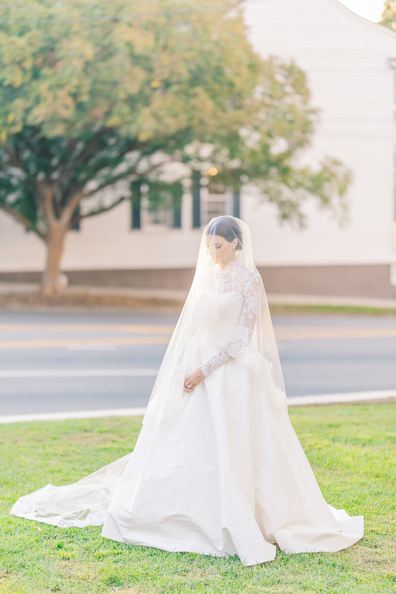 A bride in a lace gown stands in soft afternoon light, her veil cascading over her face, with a charming historic town setting behind her