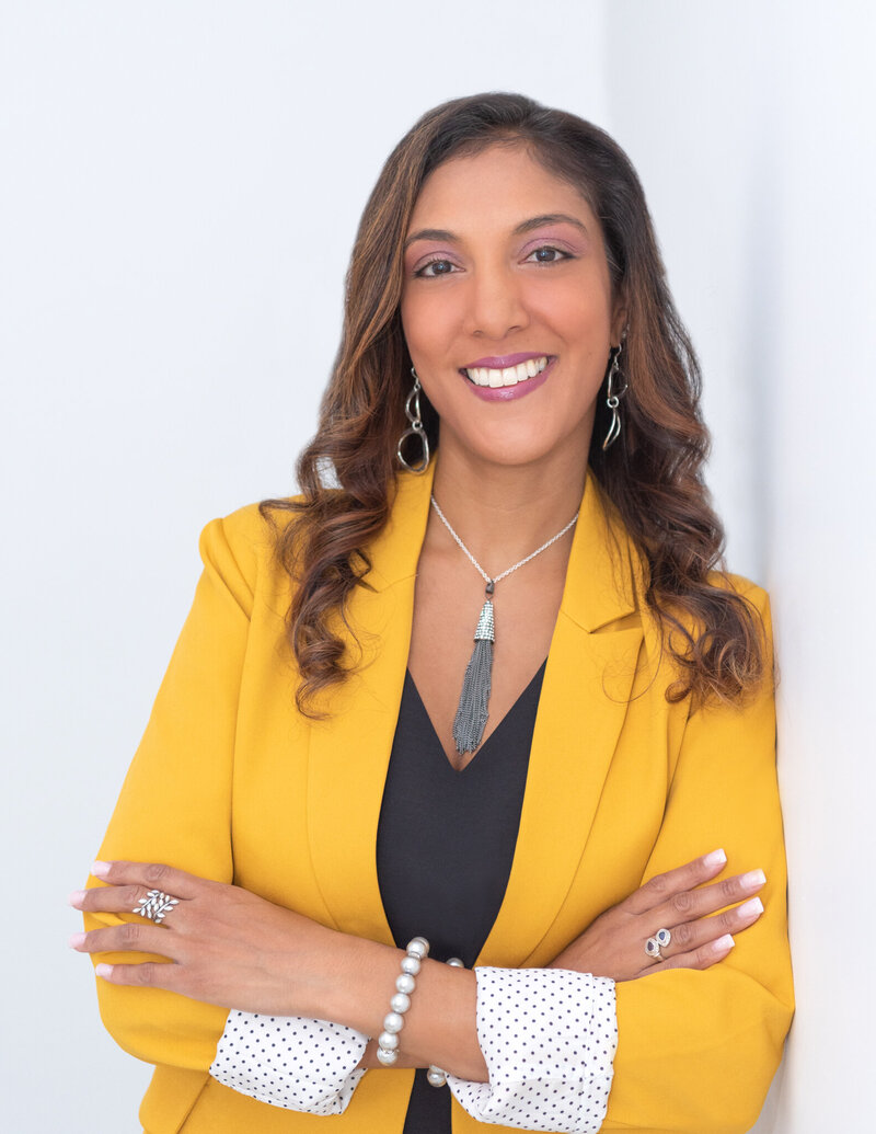 A woman wearing a yellow blazer posing with her arm crossed and leaning against a white wall for her headshots.