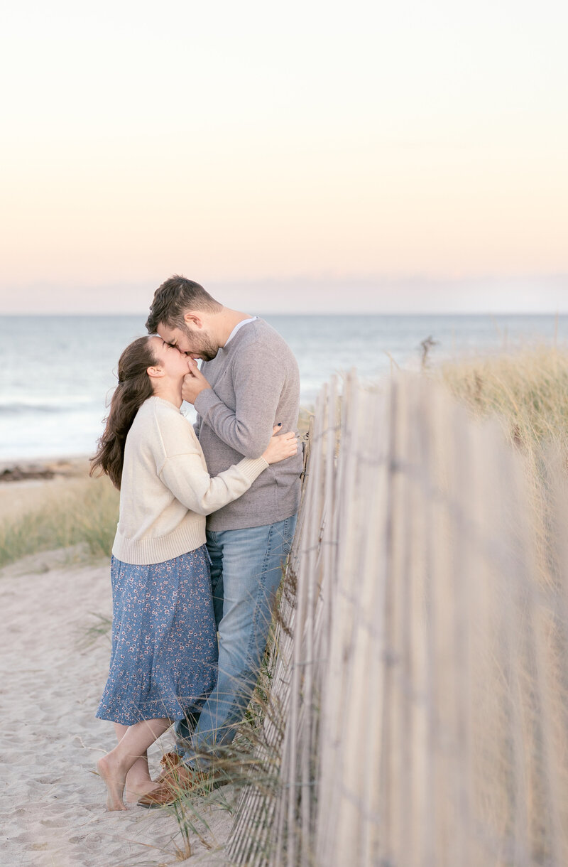 Sunset engagement photo of a couple kissing by a fence at Duxbury Beach, Massachusetts.
