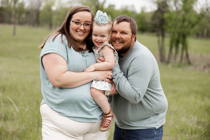 Family portrait of a mother, father and toddler daughter all standing, cuddling and smiling at the camera. 
