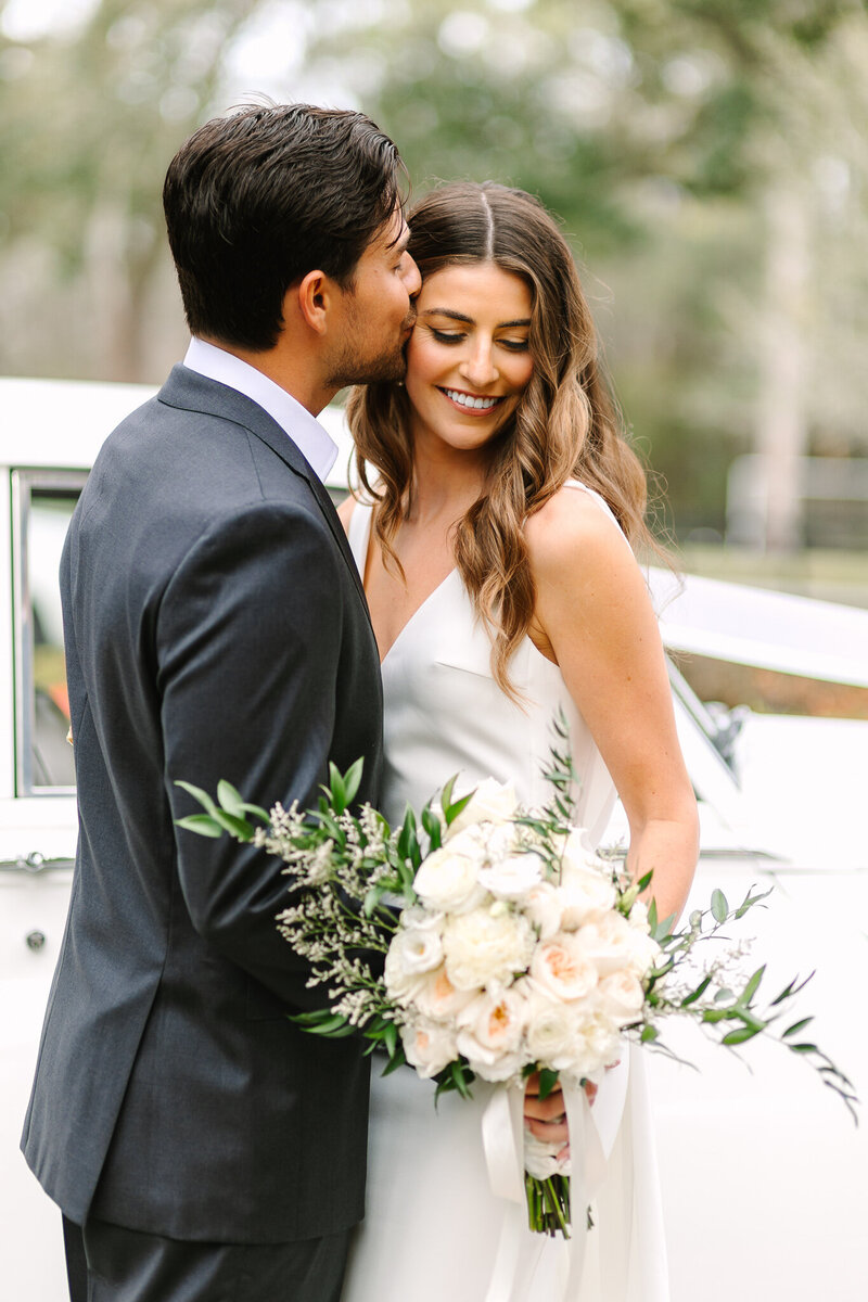 Groom kissing bride on temple as she smiles downward
