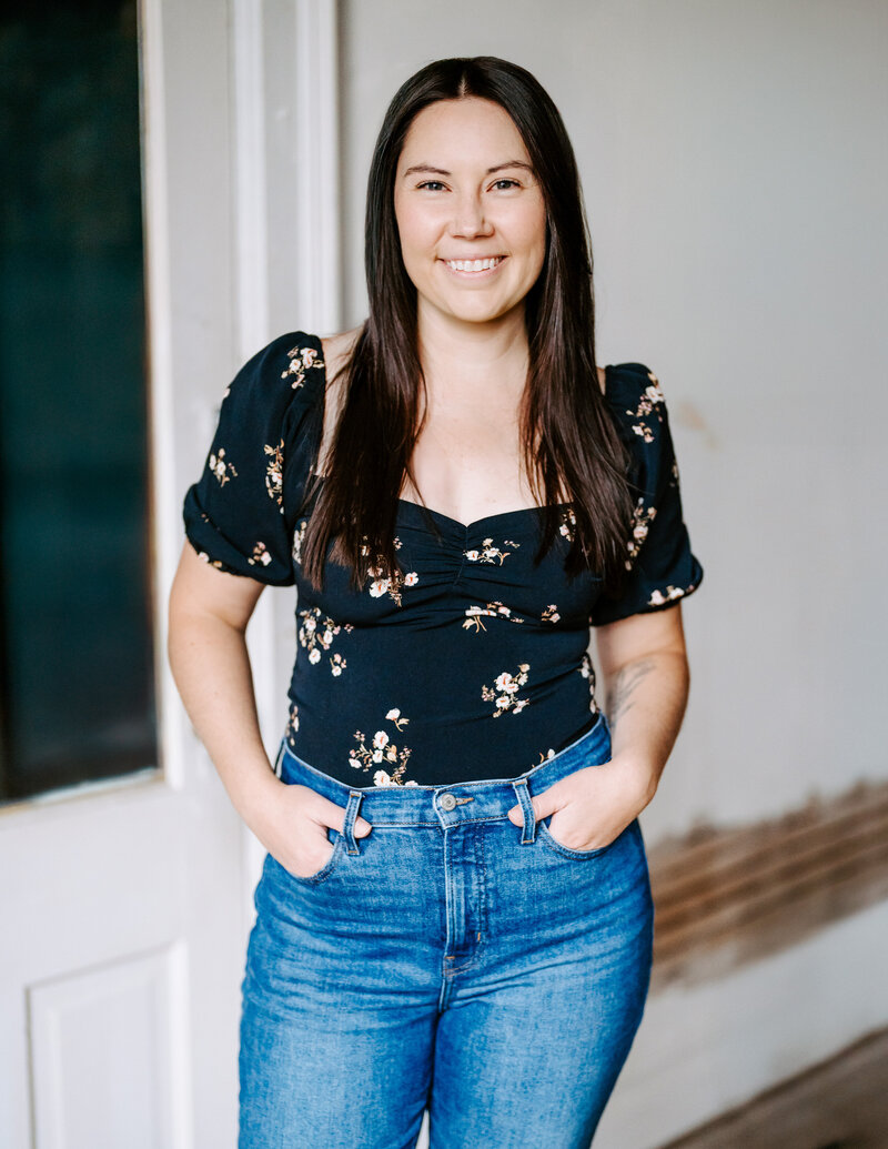 Portrait of Colorado wedding and portrait photographer Avenir Photo Co. standing with her hands in her pockets with a black floral short sleeve shirt smiling at the camera. 