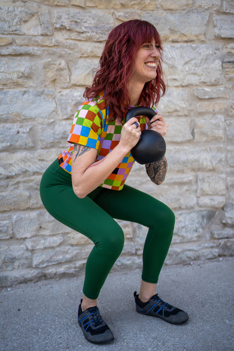 a woman in a rainbow checkered shirt does a goblet squat with a kettlebell to demonstrate online strength training programs