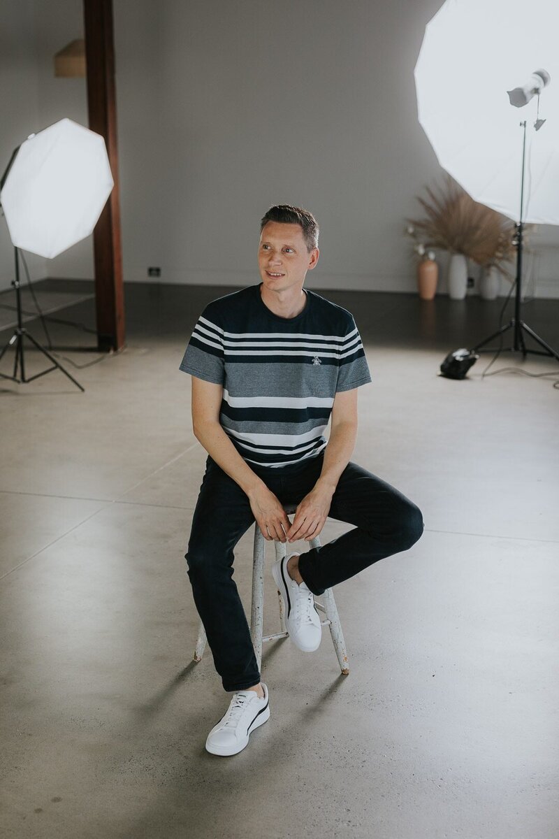 Photo of Chris Perkins, Photographer at Underatreehouse Photography, a commercial event photographer on the Central Coast. The image shows him sitting on a stool in a photography studio with studio lights in the background