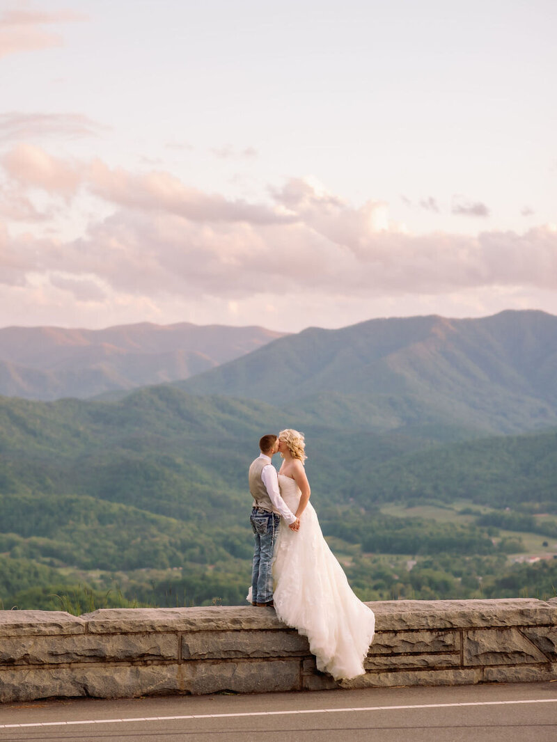 couples eloping to Gatlinburg in the Foothills with bride and groom embracing and kissing on a stone wall with the Smoky Mountains behind them photographed by photographers in gatlinburg tn