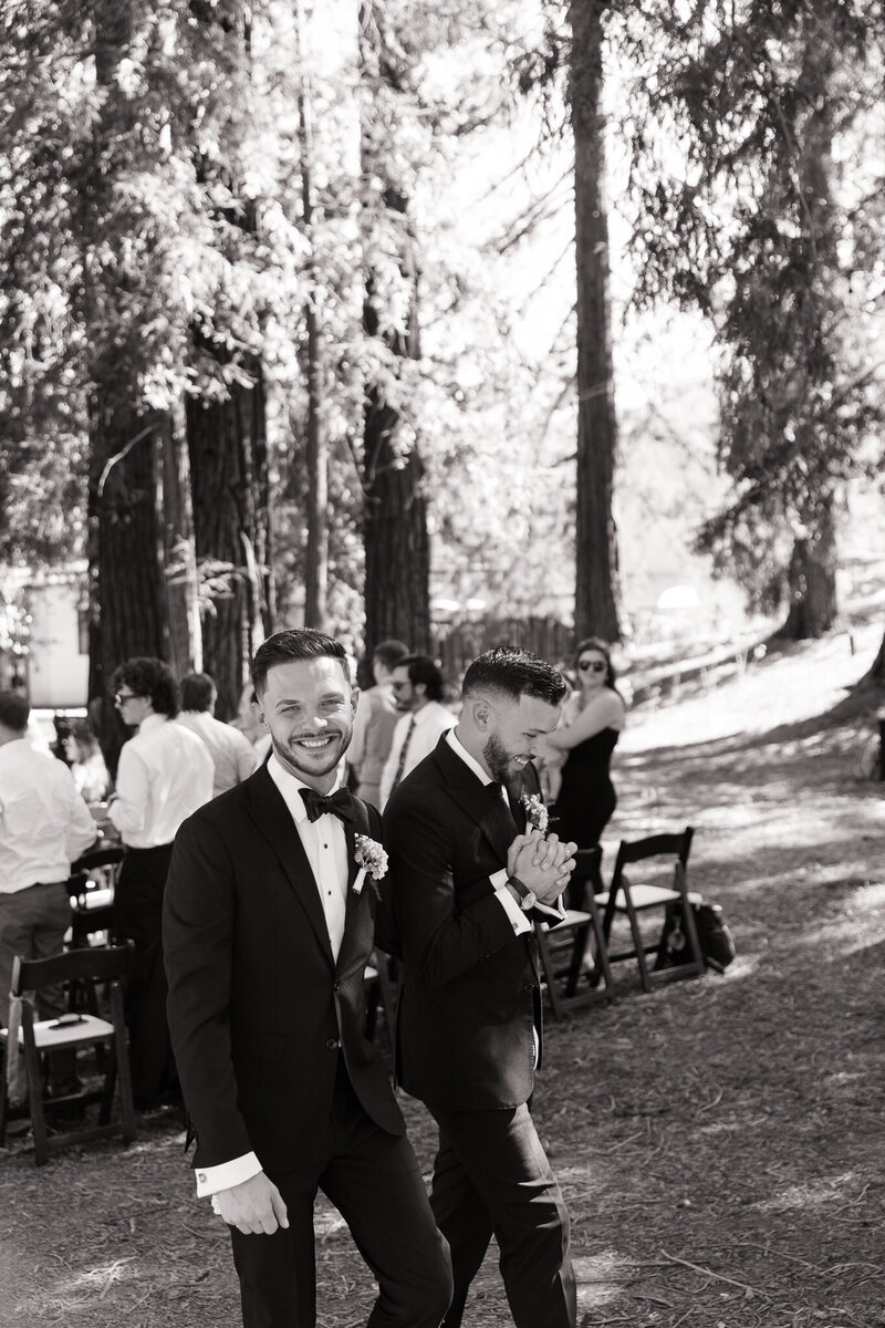 Two grooms holding hands and laughing as they walk back down the aisle together after their outdoor redwood wedding ceremony in Ukiah, California