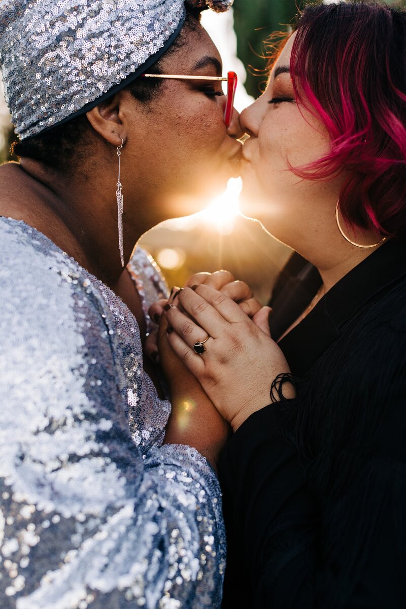 a couple kisses while holding hands with a sunflare coming between them