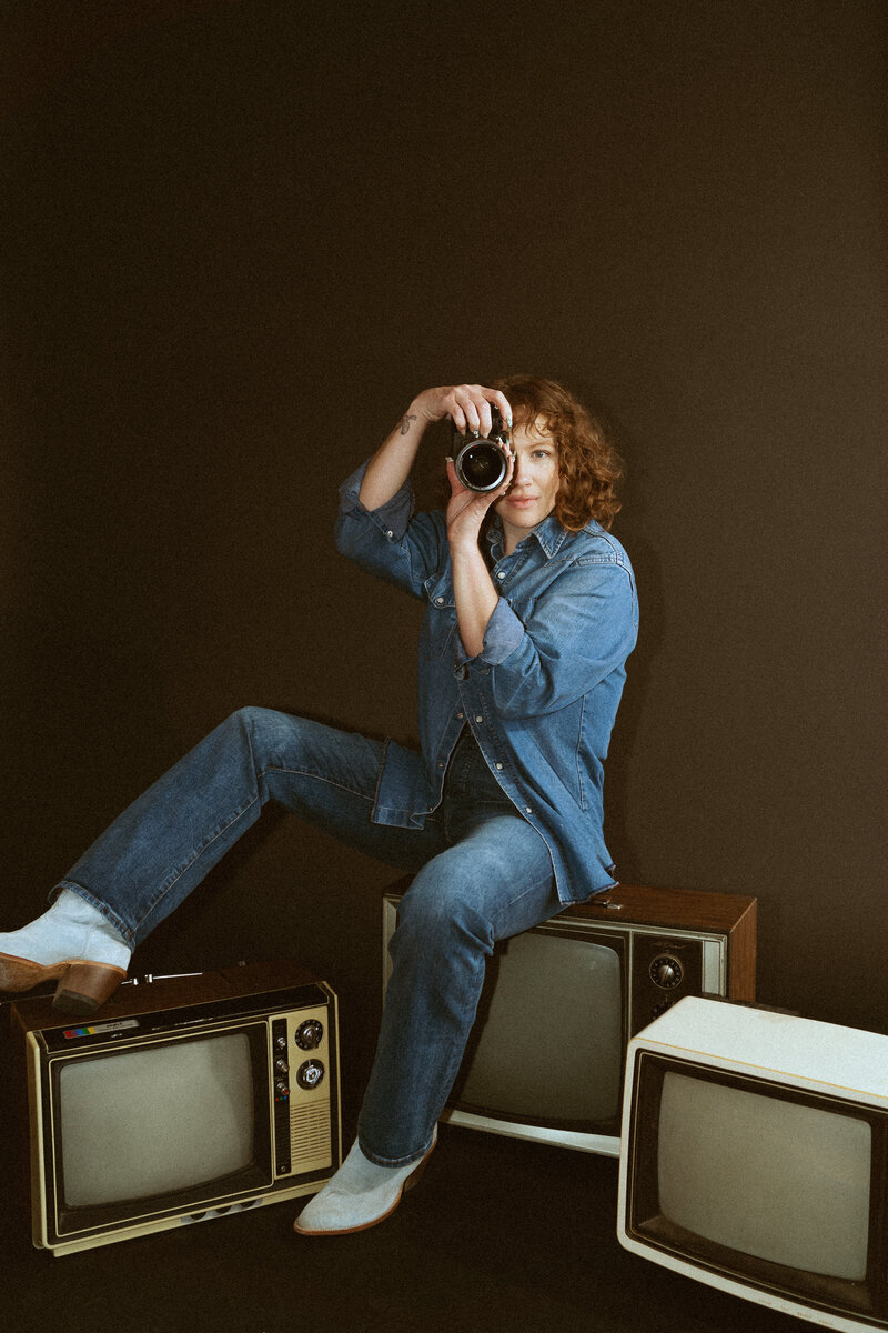 photographer sitting on vintage televisions with a camera up to her face