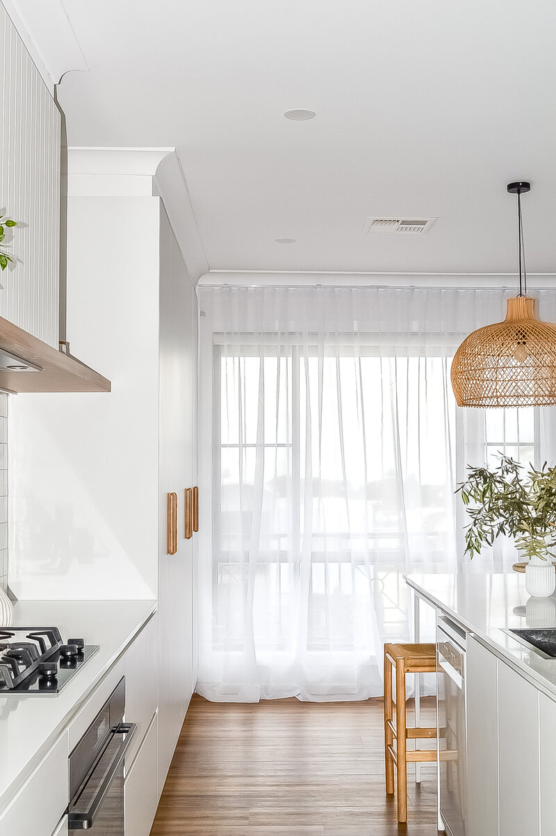 Bright kitchen with white cabinets, wooden stools, sheer curtains, and a woven pendant light—a testament to the meticulous craft of Newcastle interior design.