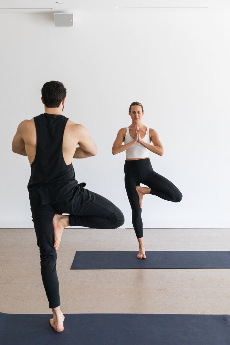 Yoga instructor guiding a male client in tree pose, both practicing balance and mindfulness together.