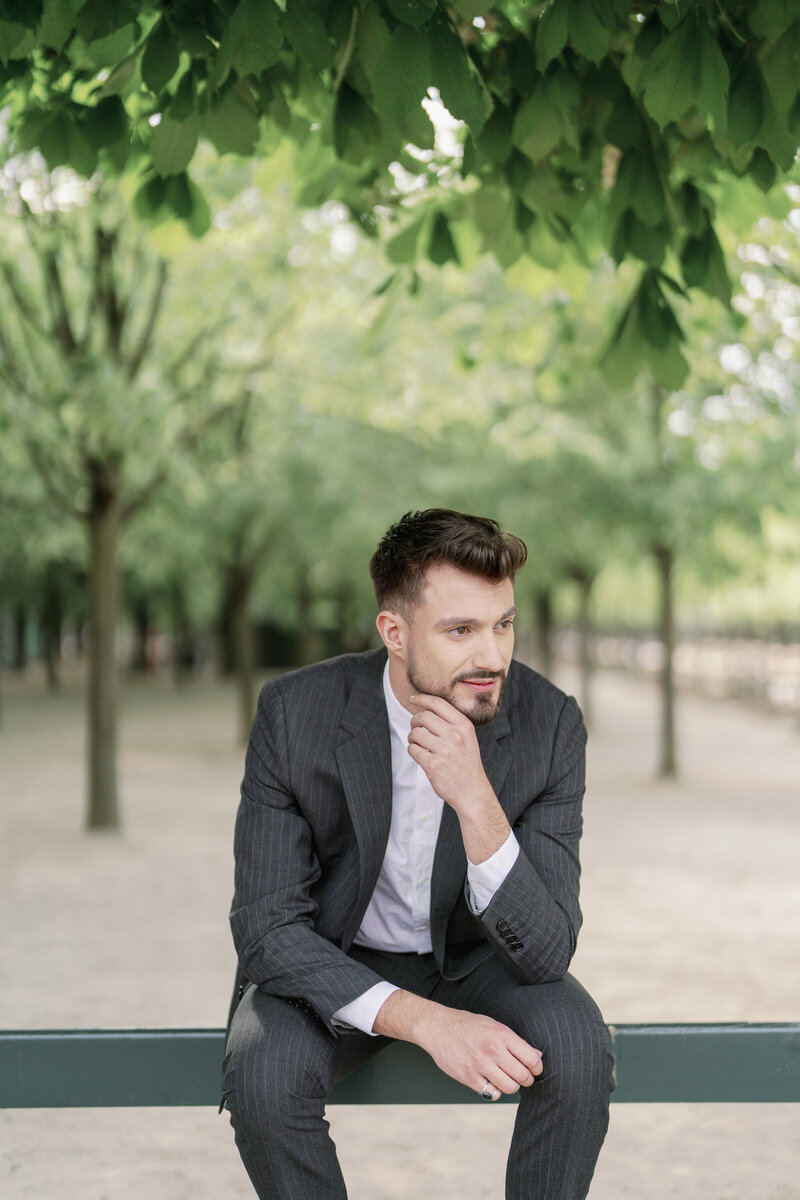 a man with dark brown hair in a dark grey suit sitting on a park bench in jardin du luxembourg looking off into the distance  and lightly touching his chin