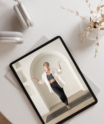 A tablet is placed on a light surface, next to a vase with dried flowers and wireless headphones. On the tablet screen, Sarah is visible, standing on a yoga mat in a spacious, bright room with an arch in the background. She appears to be demonstrating a yoga pose or part of a sequence, captured in a moment of instruction or practice. This setup suggests a peaceful, focused environment for learning and practicing yoga remotely
