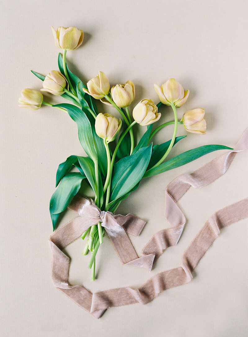 A hand-tied bouquet of pale yellow tulips with green leaves, styled on a neutral background with a soft taupe ribbon artfully draped beside it.