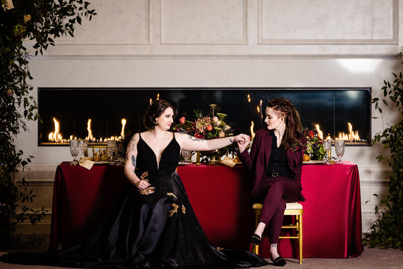 Bride in maroon suit holding bride's hand in black dress