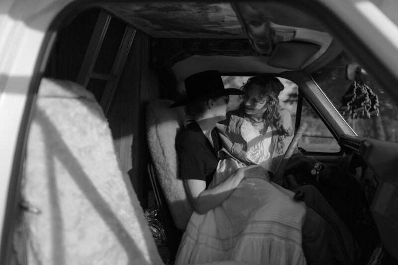 black and white photo of a couple sitting in the front seat of a vintage camper van, sharing an intimate and playful moment. The woman, dressed in a flowing, bohemian-style white dress, sits sideways on her partner’s lap, leaning in with a joyful smile. The man, wearing a wide-brimmed hat and dark t-shirt, gazes up at her with a look of affection. Soft sunlight streams through the van windows, casting gentle shadows across the cozy interior, which features a sheepskin seat cover and rustic decor. 