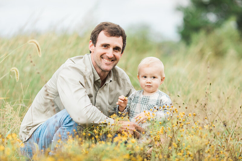 dad kneels down next to his 2 year old son in a field with yellow flowers and smiles