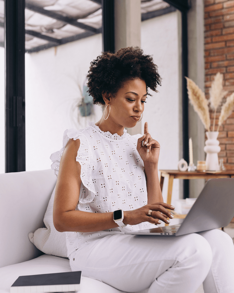 A woman wearing a white eyelet top and hoop earrings sits on a white couch, focused on her laptop. She raises a finger thoughtfully, suggesting deep focus.
