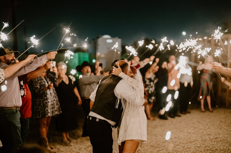 A couple shares a kiss amidst a festive scene with guests holding sparklers around them at a night event. The woman, wearing a short white lace dress with fringe details on the sleeves, and the man in a black vest and cowboy hat, create a joyous and intimate moment under the twinkling lights of the sparklers.