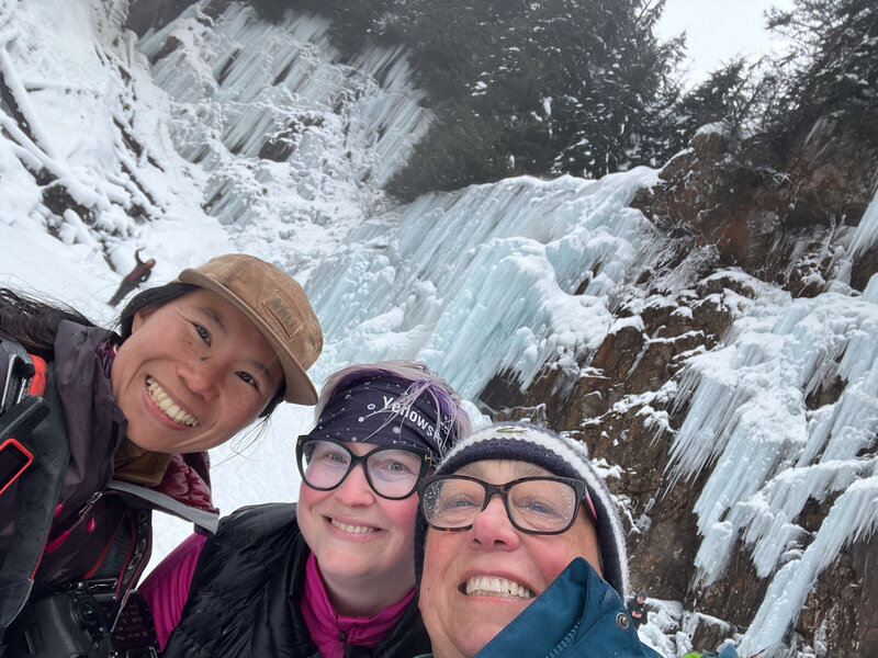 Happy brides are smiling in front of a frozen waterfall with their photographer