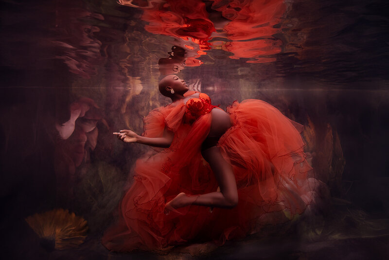 Ethereal underwater maternity photo of a dark skinned woman in a vibrant orange tulle gown floating in the water just below the surface and her reflection