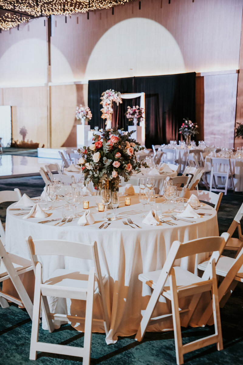 A beautifully decorated indoor wedding reception featuring round tables covered in white linens. The tables are set with elegant floral centerpieces, glasses, and candles, with a backdrop of soft lighting and an arch.