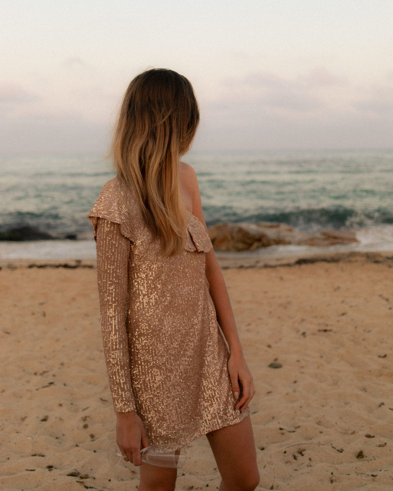 Woman in a sequin dress standing on a sandy beach, facing the ocean under a pastel sunset sky