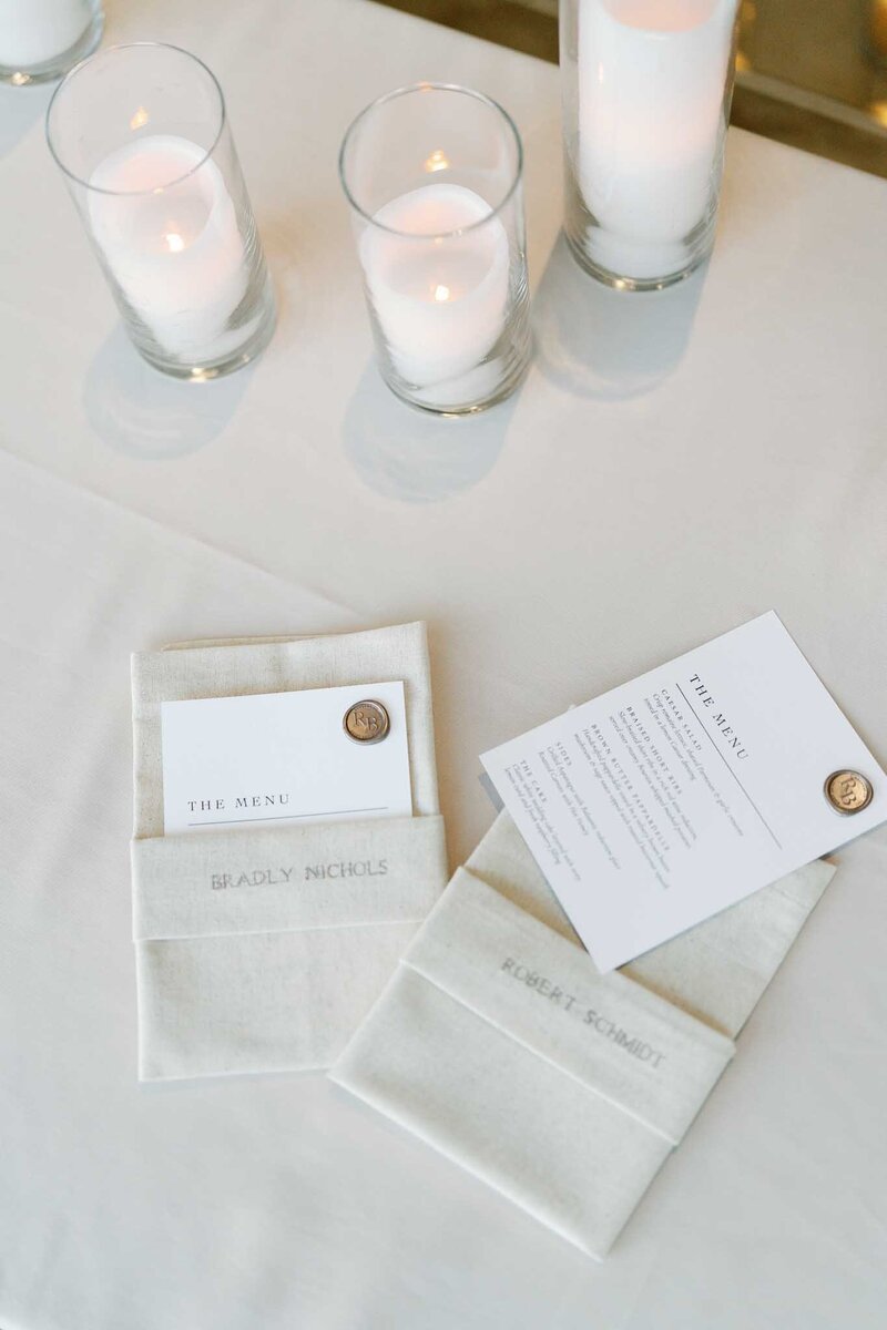 folded and embroidered linen napkins with menu cards with was seals on a white tablecloth covered table
