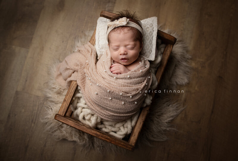 Newborn baby girl wrapped in brown wrap with pearls on it laying in a cute baby bed
