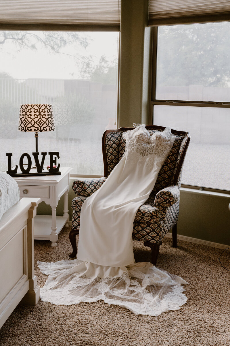 An elegant wedding dress with lace detailing and a long flowing train is draped over a vintage patterned armchair beside a window. The room is warmly lit, featuring a lamp with a decorative shade and a white nightstand with a sign spelling 'LOVE'. The tranquil domestic setting creates a romantic and serene atmosphere, perfect for bridal preparations.