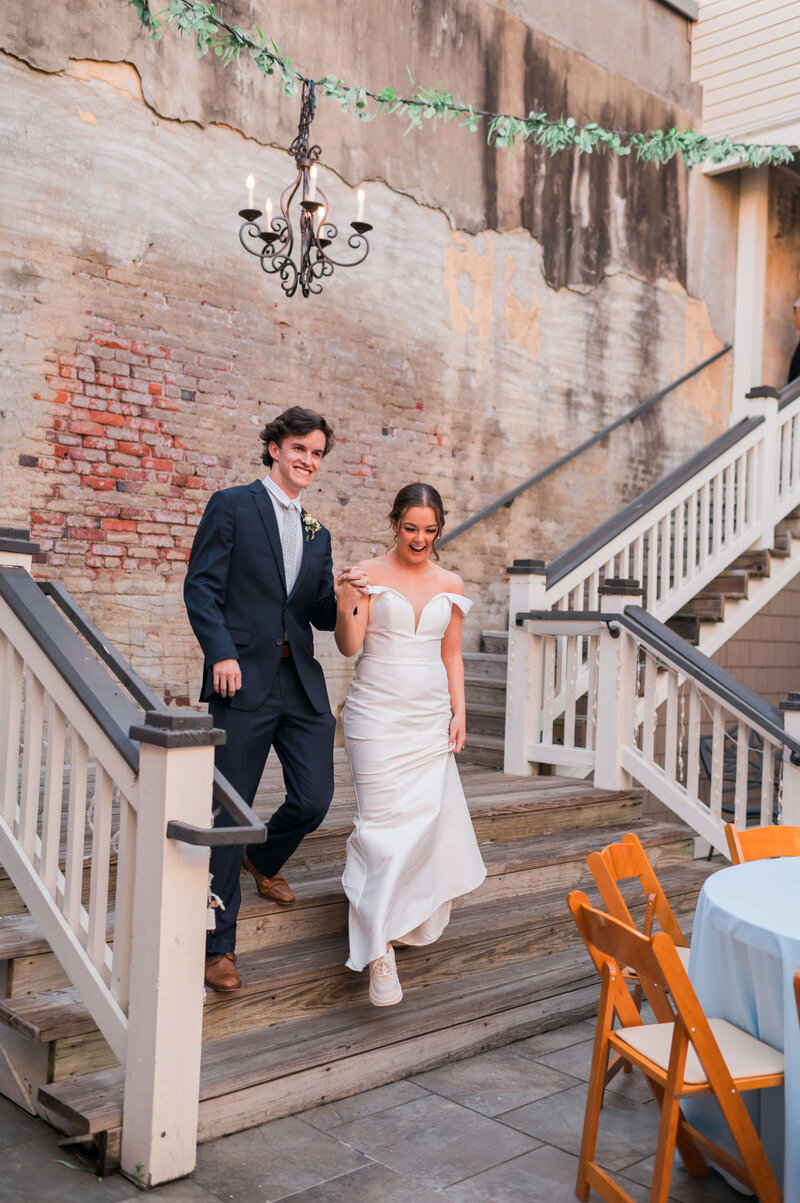 A smiling bride in a white gown and sneakers walks down outdoor steps with a groom in a navy suit. They hold hands, under a black chandelier, with exposed brick wall and wooden railings in the background.