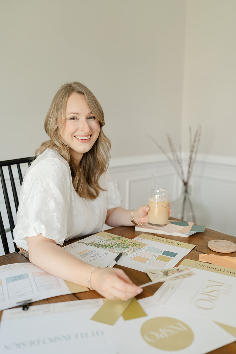 Woman sitting at a table of color swatches and brand elements holding an iced latte and smiling