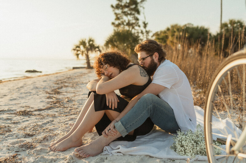 young brunette married couple snuggle intimately on picnic blanket on beach during couples portrait session with Brittney Stanley