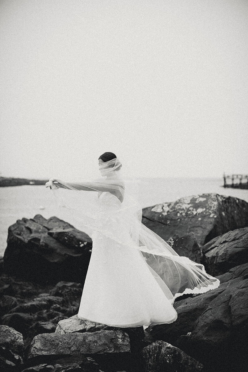 Bride with flowing veil standing on coastal rocks overlooking the ocean during her Maine wedding.