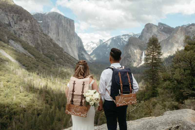 Adventure Elopement Ideas | Eloping couple stands in front of a huge mountain view with hiking backpacks that say "Just Married"