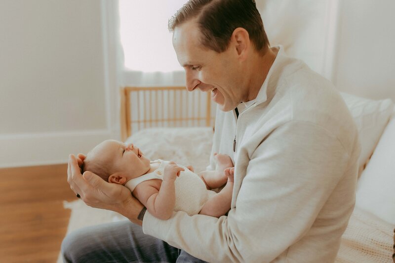 A man in a light sweater holds a baby while sitting on a bed in a softly lit room.