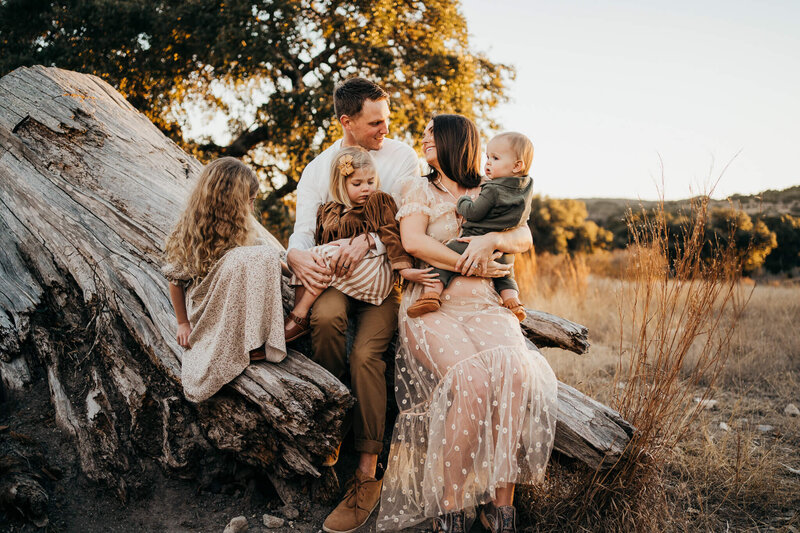 A family of parents and three little girls sitting on a log  and smiling at each other
