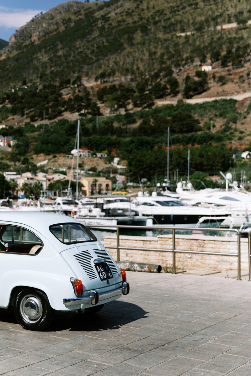 Blue Fiat parked beside yachts at an Italian marina, evoking the relaxed sophistication of luxury travel.