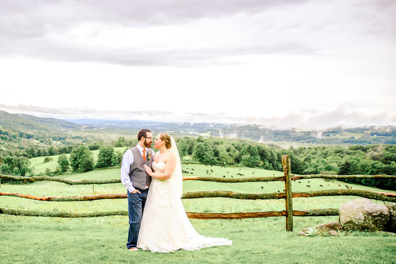 A bride and groom pose in front of a split rail fence overlooking  a gorgeous countryside