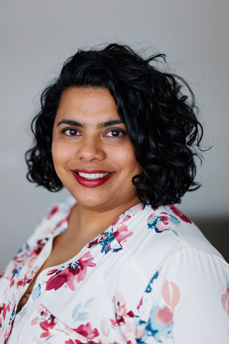 Portrait of a woman with dark, curly hair, wearing a white floral blouse and smiling warmly at the camera.
