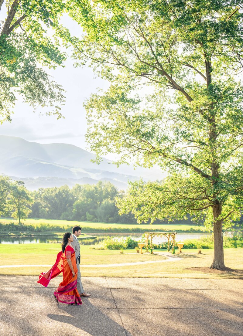 A couple in traditional attire walks along a path under a large tree. The woman wears a red sari, and the man is in a light-colored suit. They are in a sunlit park with mountains and a small decorated stage in the background.