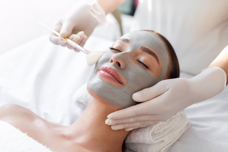 Closeup image of a woman lying down getting a green facial treatment from an esthetician wearing white gloves