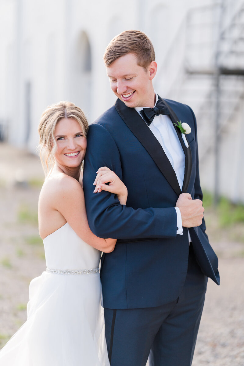 A bride in a white dress hugs onto her husband's arm and smiles while he looks down and smiles at her