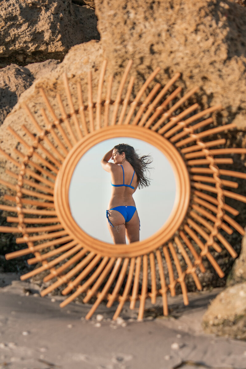 Model can be seen in reflection of rattan mirror wearing blue recycled fabric bikini