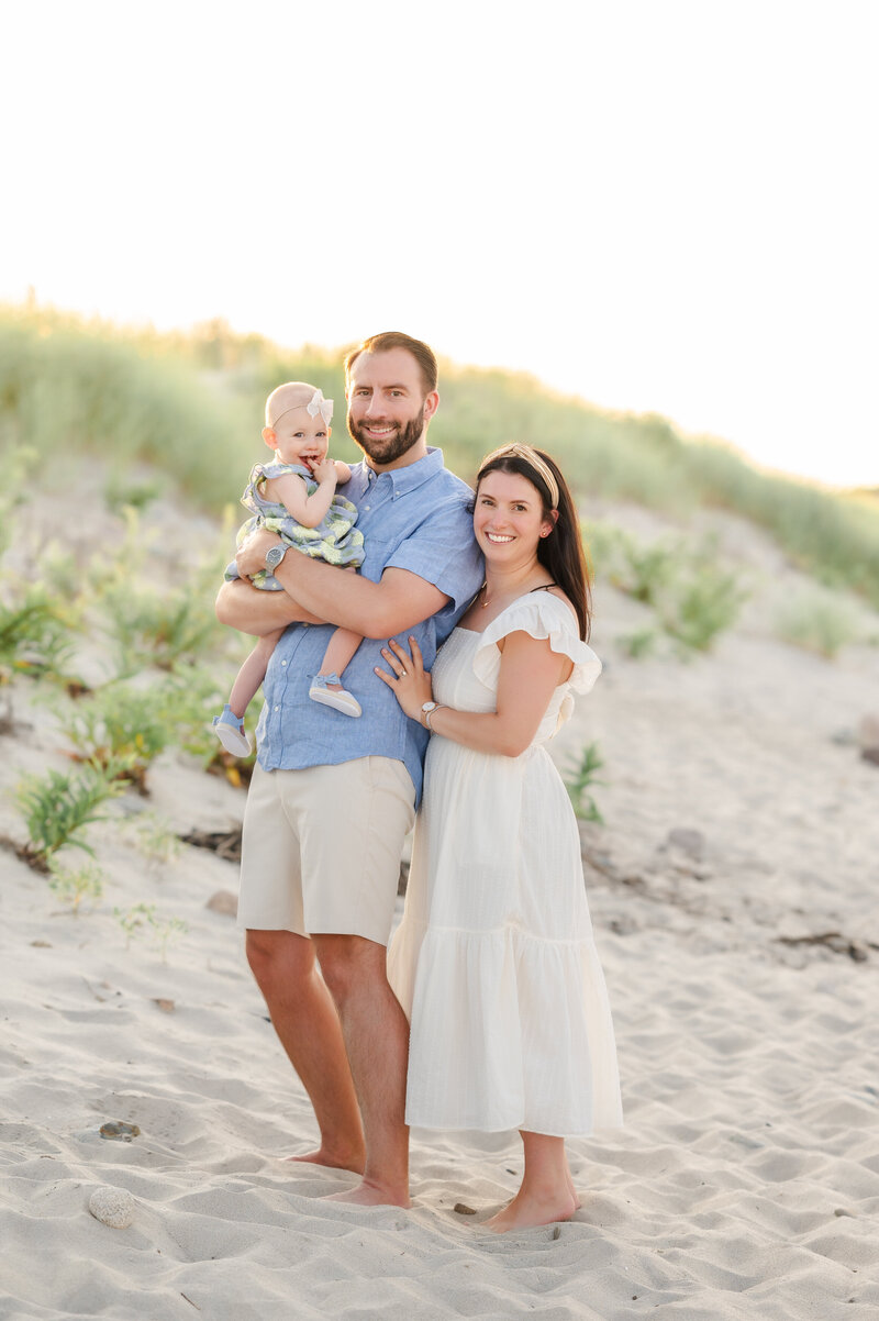 dad holding baby while standing on the beach with mom standing next to them and smiling