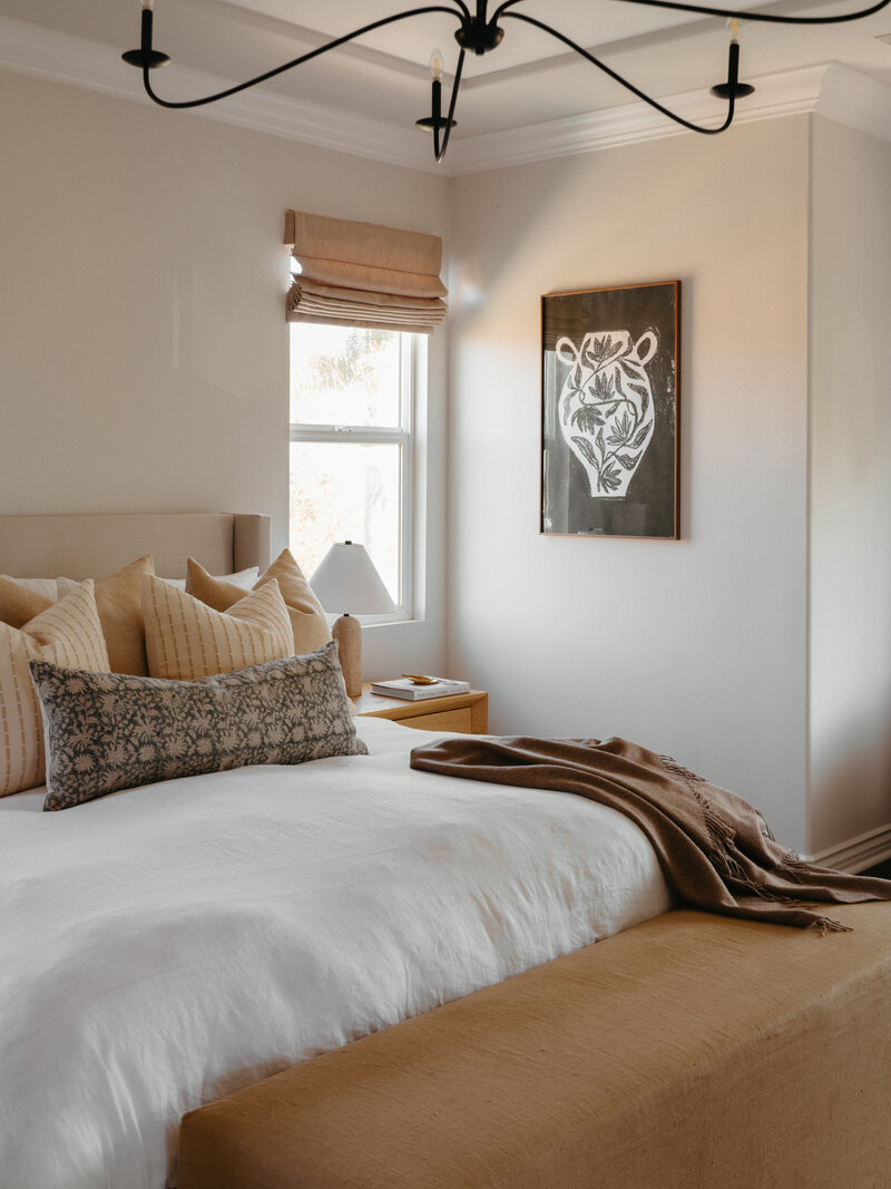 Warm and airy San Diego bedroom featuring neutral layered bedding, natural oak nightstand, rustic wood console table with curated styling, and a black sculptural chandelier — a perfect blend of California casual and organic modern design.