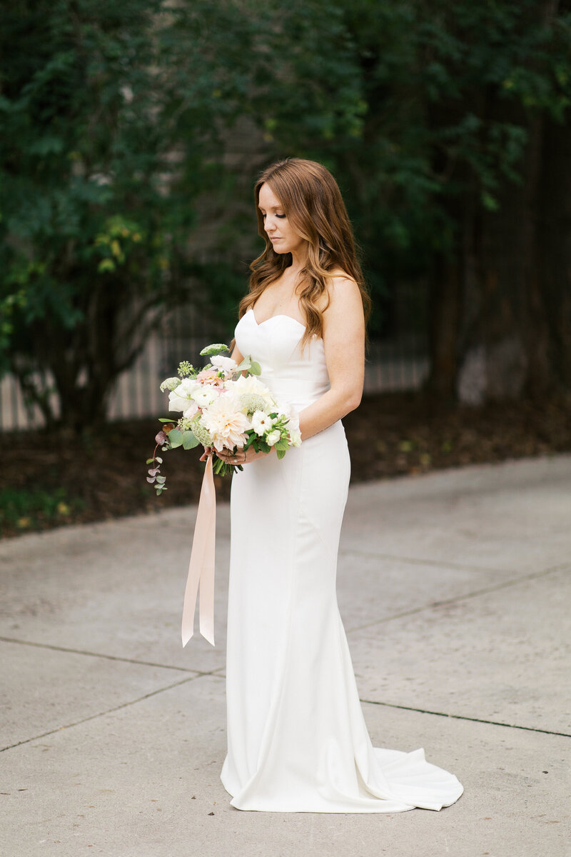 Bride in ivory wedding dress standing tall and looking at her beautiful ivory bouquet at St. Paul College Club wedding. Captured by Minnesota wedding photographer.