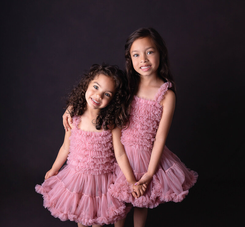 Sisters standing in top Philadelphia family photo studio for siblings portrait. Big sister, age 8 has her arm wrapped around her little sister, age 5. The girls forearms are holding hands. Both of the girls are smiling at the camera.