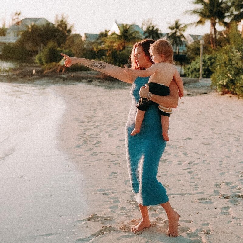 A mother is holding her child on the beach and both of them are pointing to the ocean