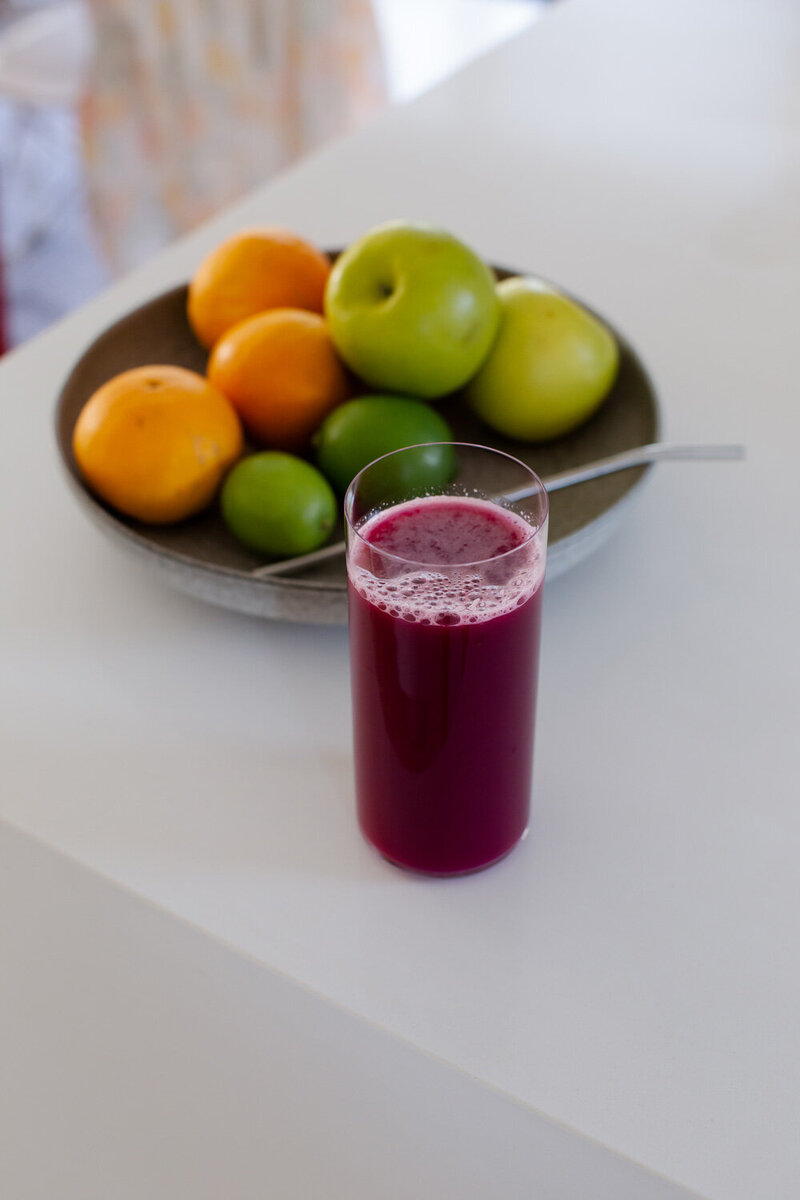 Freshly juiced beet and carrot juice in a tall glass, in front of a fruit bowl on a marble kitchen bench.