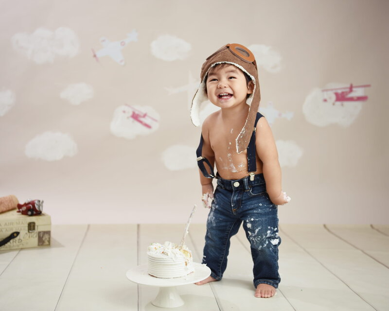 A baby boy dressed as a pilot in suspenders and a hat, standing next to a cake in an airplane-themed cake smash setup with clouds and airplanes in the background.